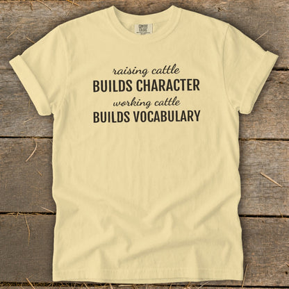 Light yellow t-shirt with black text: "raising cattle BUILDS CHARACTER, working cattle BUILDS VOCABULARY."