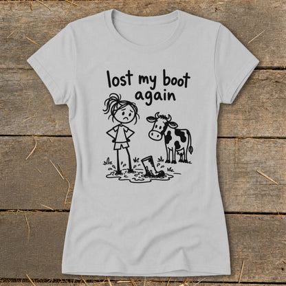 Light gray short-sleeved t-shirt with a graphic of a sad stick-figure girl and a cow looking at a muddy boot, with text reading "lost my boot again".