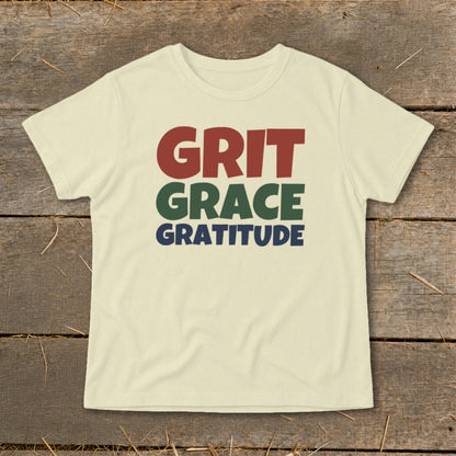 A cream-colored short-sleeved t-shirt with the words Grit, Grace, and Gratitude printed in red, green, and blue.