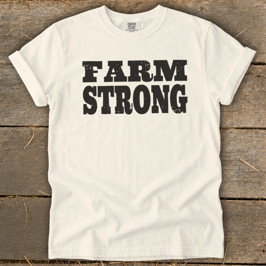 Off-white t-shirt with rolled sleeves laid flat on a weathered wooden background, featuring bold black distressed text that reads FARM STRONG.