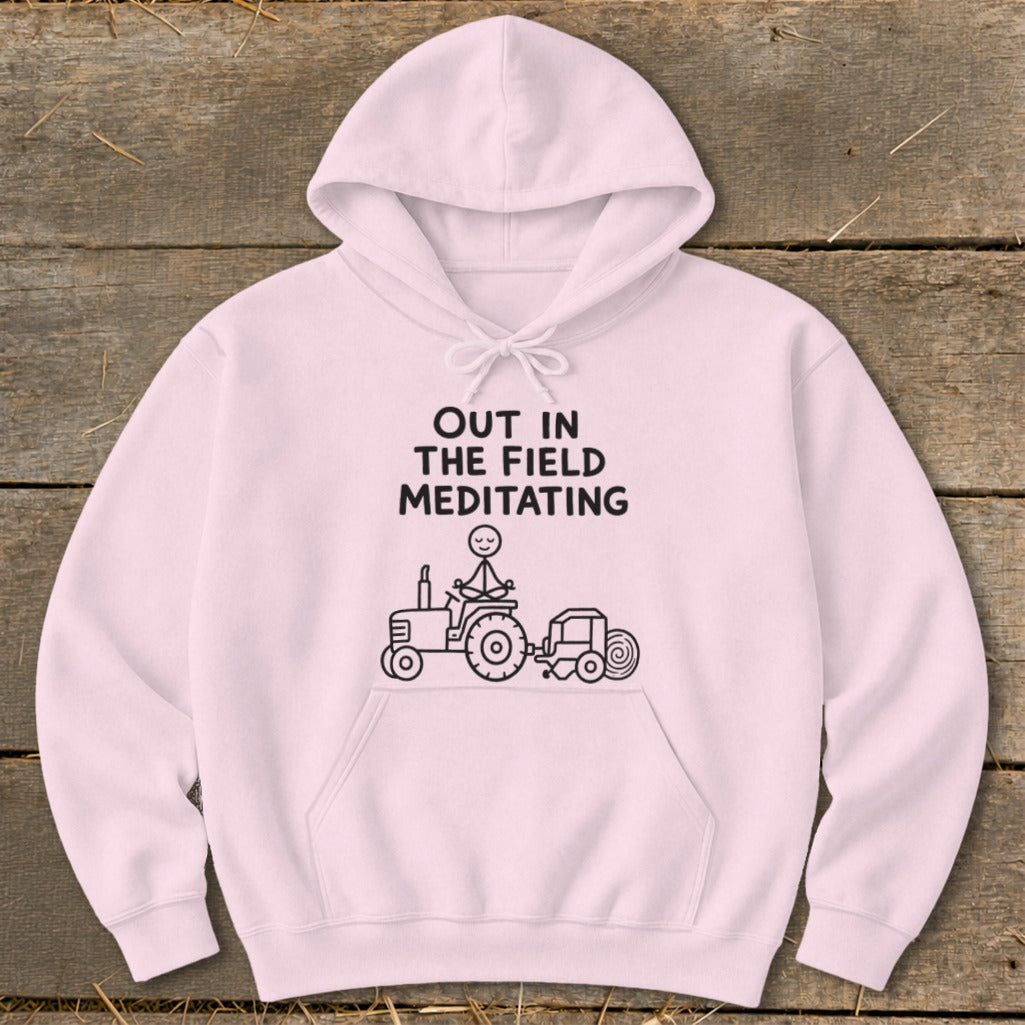 Pink hoodie with text "OUT IN THE FIELD MEDITATING" and a stick figure meditating on a tractor.