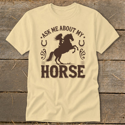 Light yellow t-shirt with brown graphic of a woman riding a bucking horse and text "ASK ME ABOUT MY HORSE".