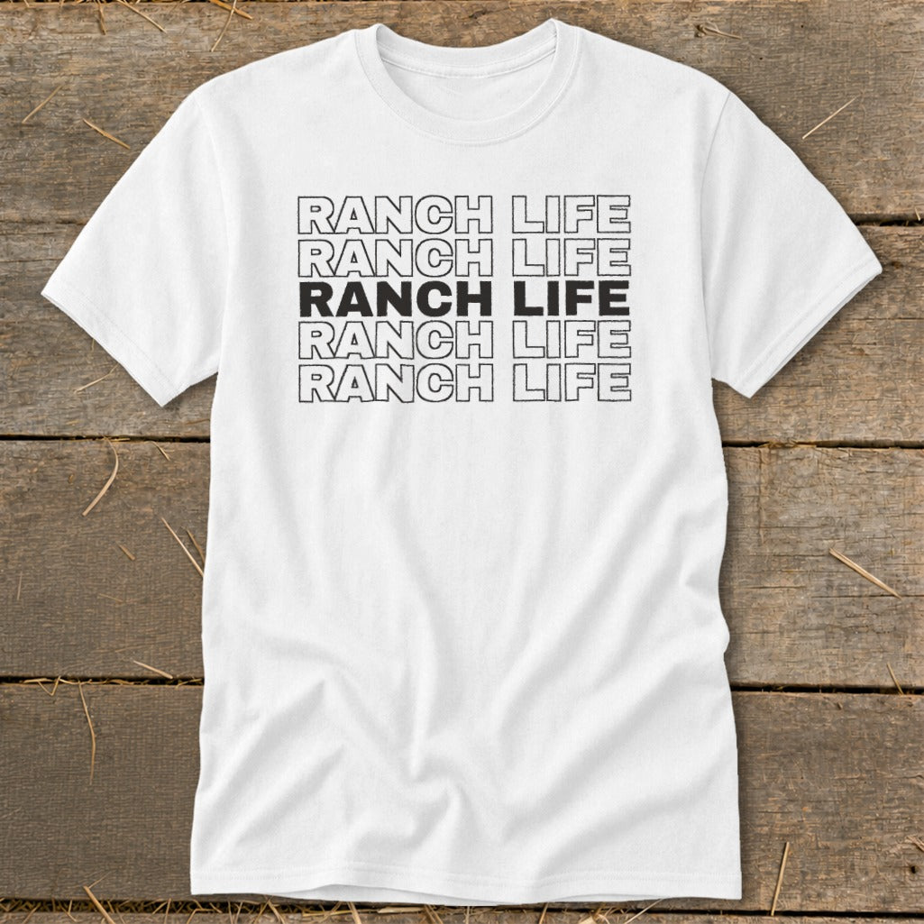 A white t-shirt featuring the text "RANCH LIFE" stacked in five rows with the middle row filled in solid black, laid flat on a rustic wooden background.