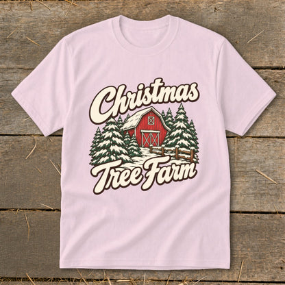 Light pink t-shirt with a Christmas tree farm graphic featuring a red barn and snow.