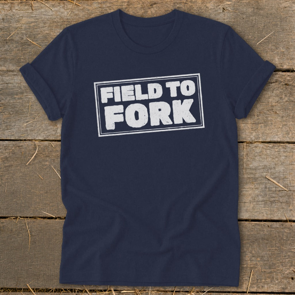 A navy blue t-shirt with rolled sleeves lies flat on a rustic wooden surface, displaying the white text "FIELD TO FORK" enclosed in a rectangular box on the chest.