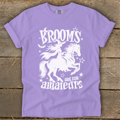 Purple t-shirt with white graphic of a witch on a horse and text "Brooms are for amateurs."