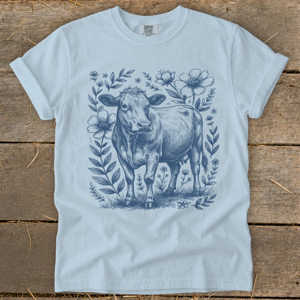 Blue line drawing of a cow surrounded by flowers on a light blue t-shirt.