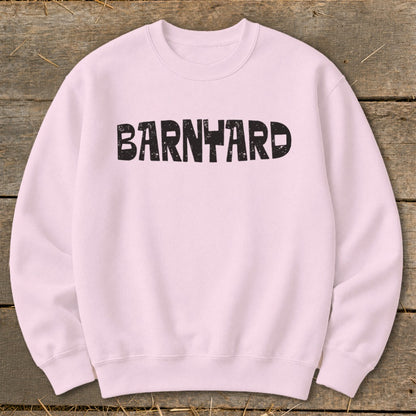 A light pink crewneck sweatshirt with the distressed black text BARNYARD centered on the chest, laid flat on a weathered wooden surface.