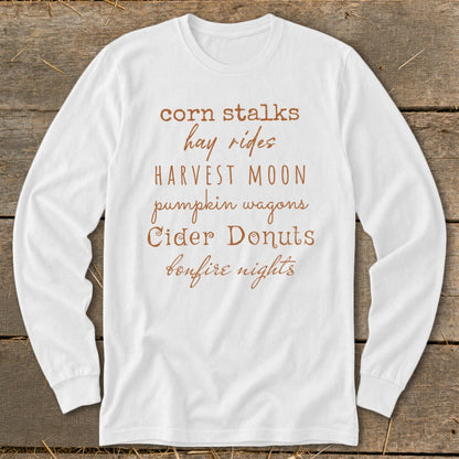White long-sleeve shirt with fall phrases: corn stalks, hay rides, harvest moon, pumpkin wagons, cider donuts, bonfire nights.