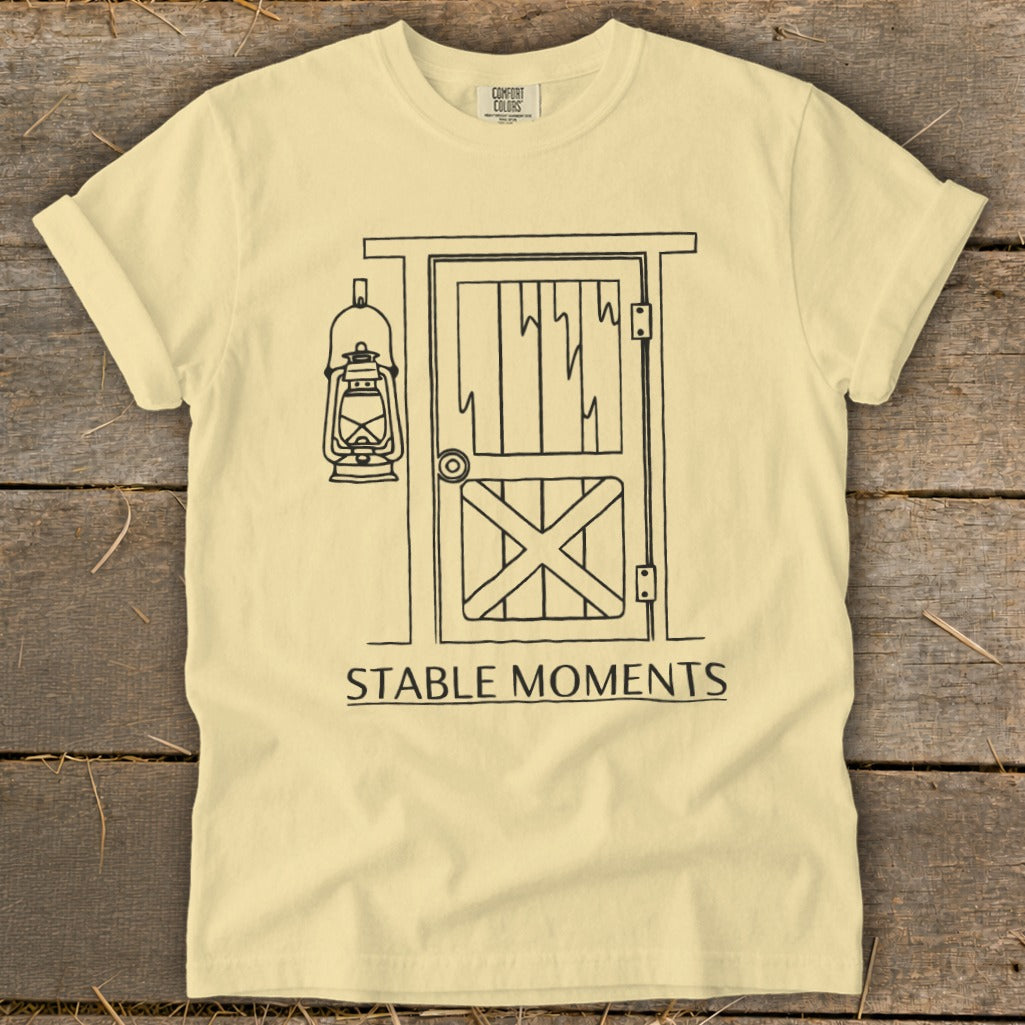 Pale yellow t-shirt with barn door, lantern illustration, and text "STABLE MOMENTS".