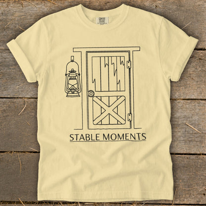 Pale yellow t-shirt with barn door, lantern illustration, and text "STABLE MOMENTS".