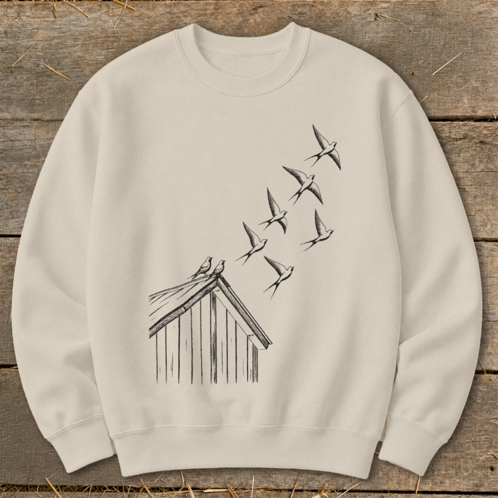 Beige crewneck sweatshirt featuring a black ink illustration of birds taking flight from a cabin roof.