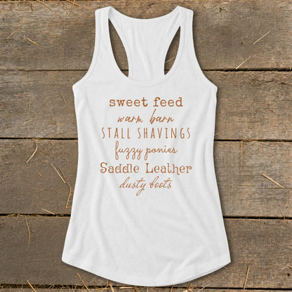 White tank top with text: sweet feed, warm barn, stall shavings, fuzzy ponies, saddle leather, dusty boots.