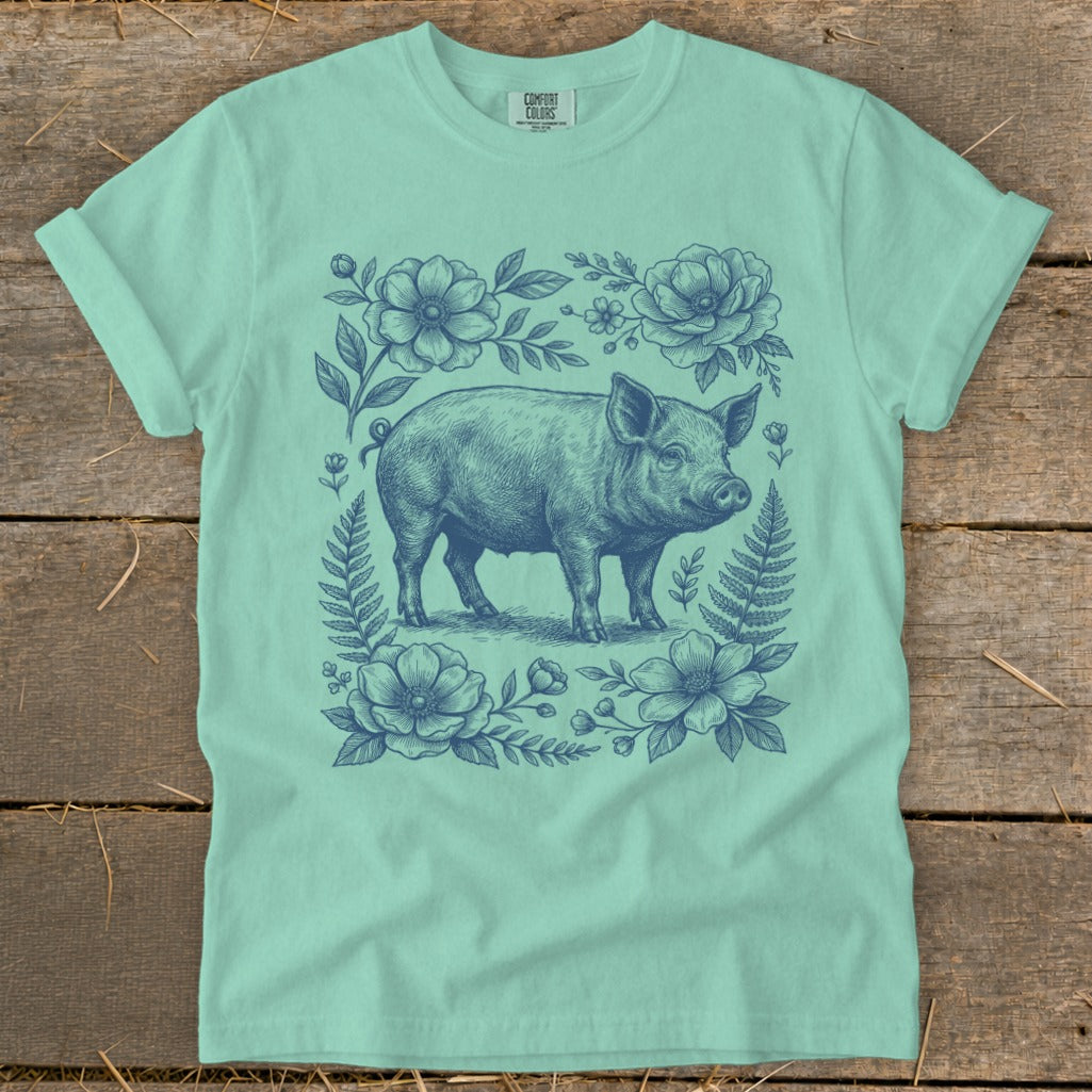 Pig graphic surrounded by floral and fern illustration on a mint green t-shirt.