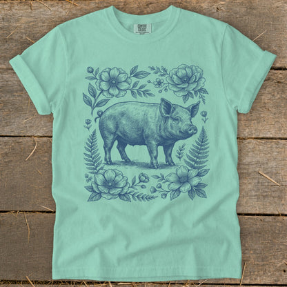 Pig graphic surrounded by floral and fern illustration on a mint green t-shirt.