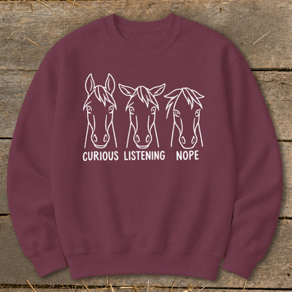 Maroon crewneck sweatshirt featuring white line art of three horse heads with the text "CURIOUS," "LISTENING," and "NOPE."