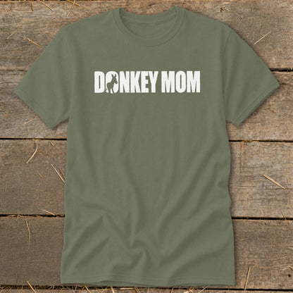 An olive green t-shirt featuring the white text DONKEY MOM, with a donkey silhouette inside the letter O, laid flat on a rustic wooden background.