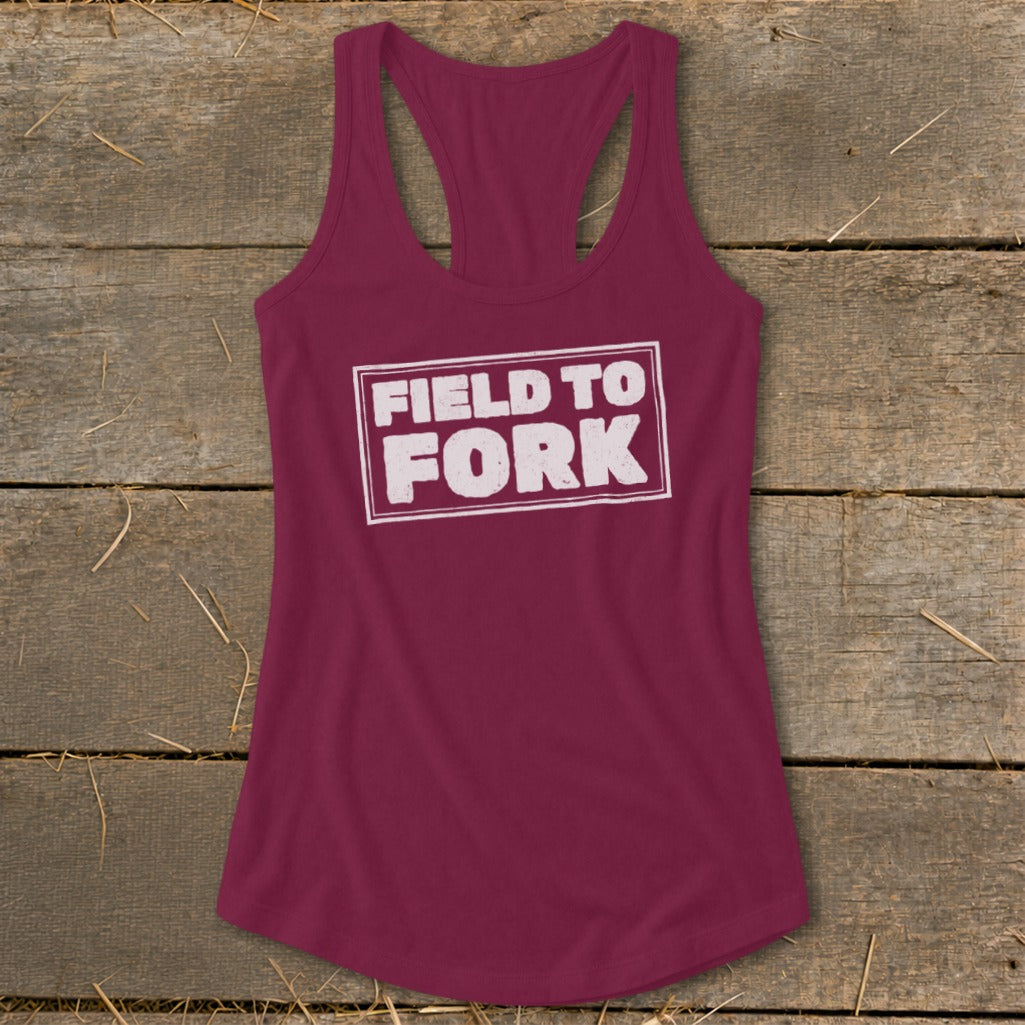 Maroon racerback tank top with the white text "FIELD TO FORK" inside a rectangular box, laid flat on a rustic wooden surface.