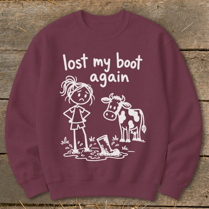 Maroon crewneck sweatshirt featuring white text that reads lost my boot again, alongside a drawing of a girl and a cow standing near a muddy boot.