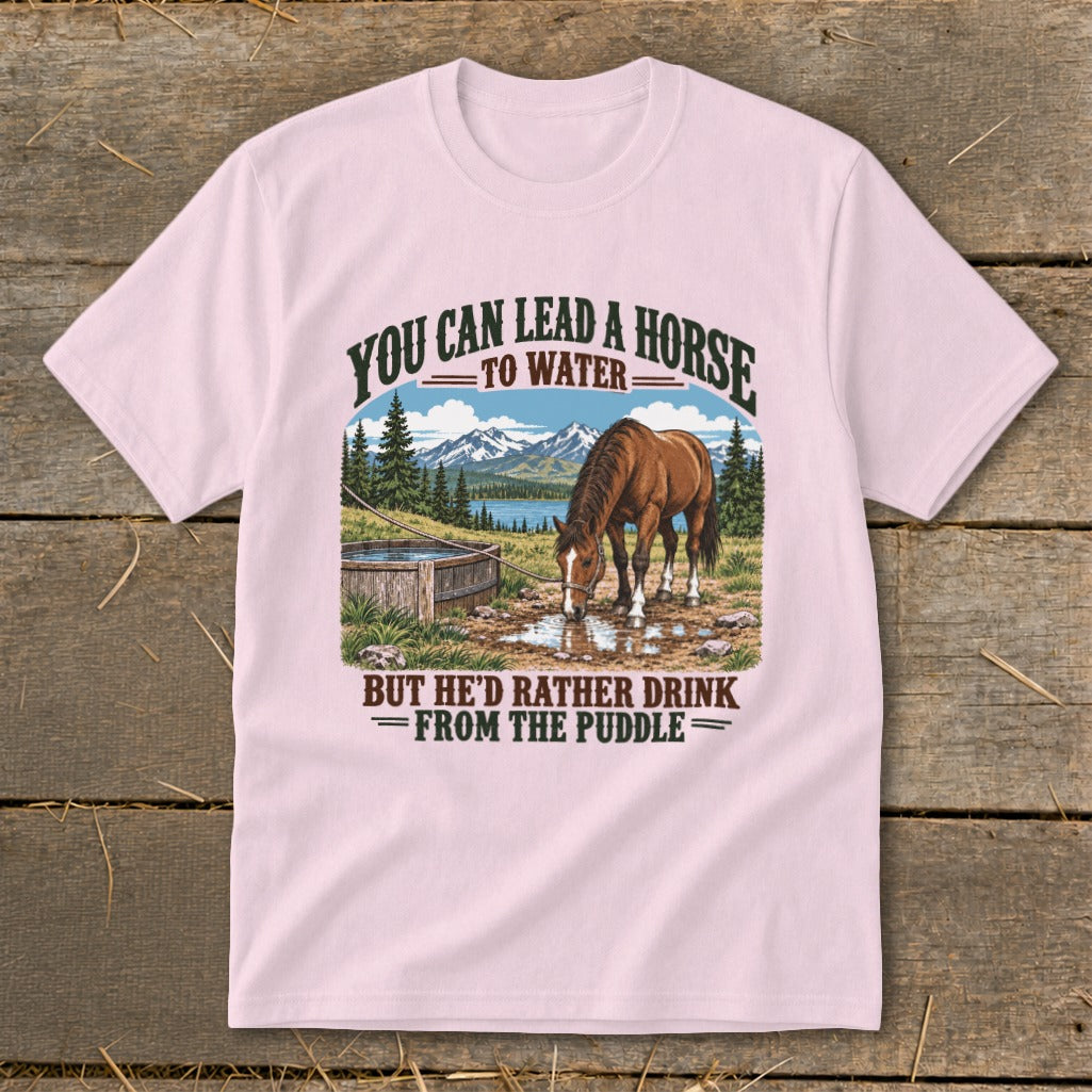 Light pink t-shirt featuring a horse drinking from a puddle with mountains and the text You Can Lead A Horse To Water But Hed Rather Drink From The Puddle.