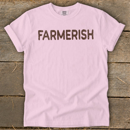 Pink t-shirt with "FARMMERISH" text in brown, laid on a wooden surface.