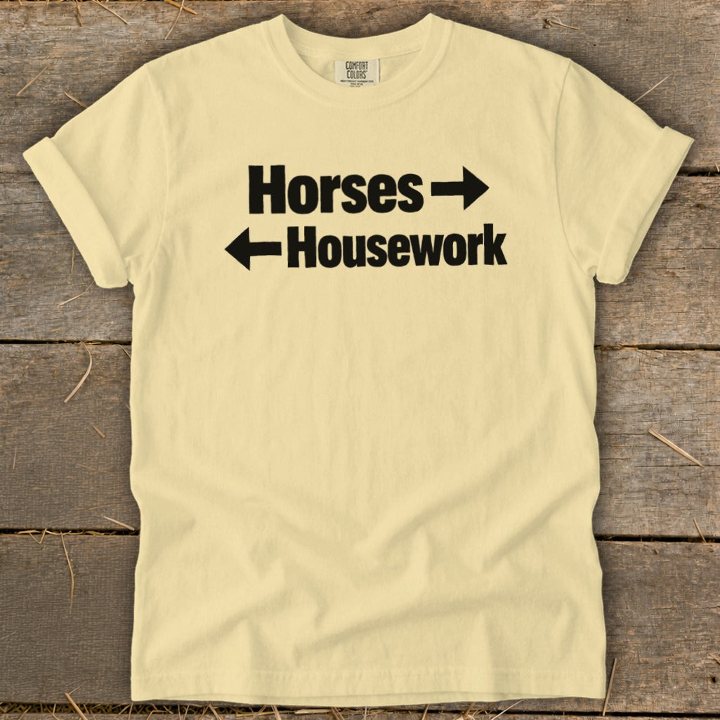 A light yellow t-shirt with black text reading "Horses" with a right-pointing arrow and "Housework" with a left-pointing arrow, laid flat on a rustic wooden background.