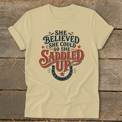 Light yellow t-shirt with "She believed she could so she saddled up" text and horseshoe graphic.