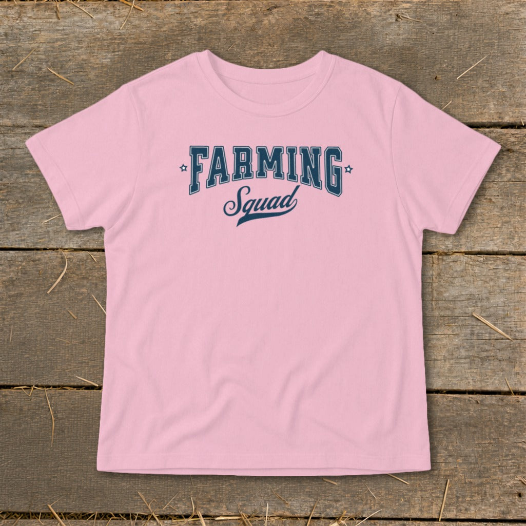 Pink t-shirt with "Farming Squad" text in dark blue on a wooden background.