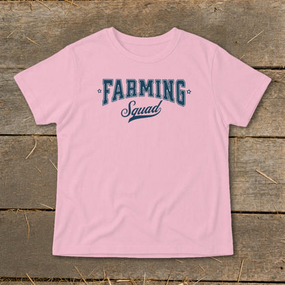 Pink t-shirt with "Farming Squad" text in dark blue on a wooden background.