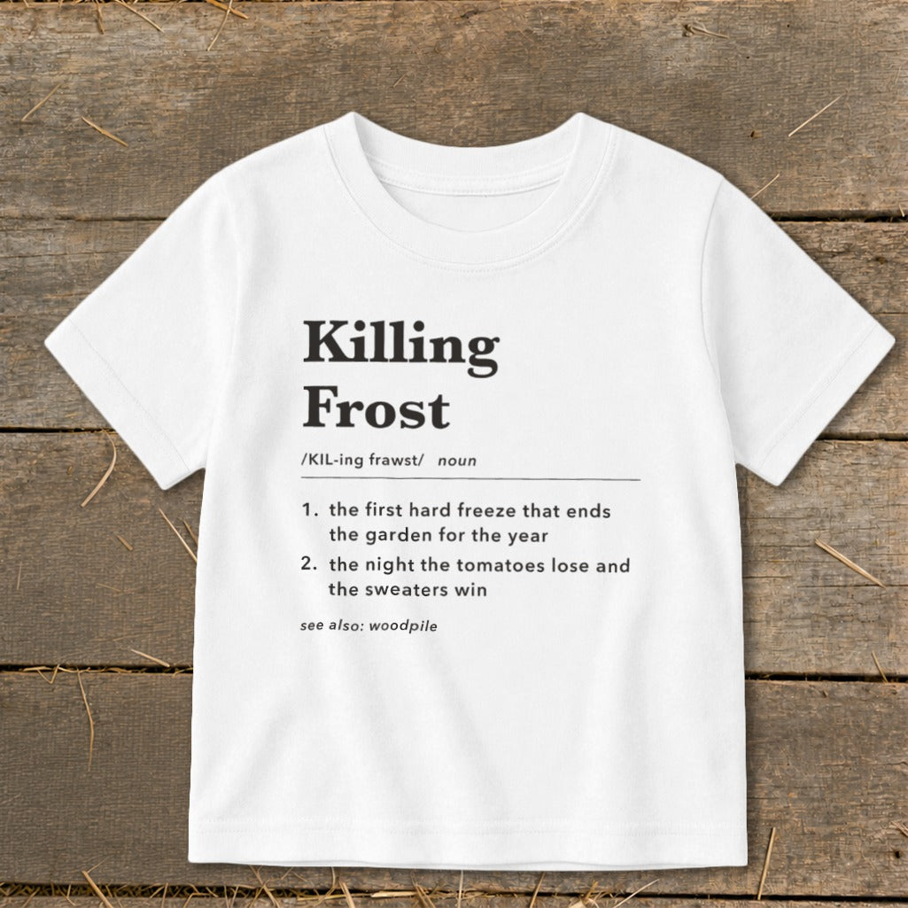 A white t-shirt featuring dictionary-style text that defines Killing Frost laid flat on wooden boards.