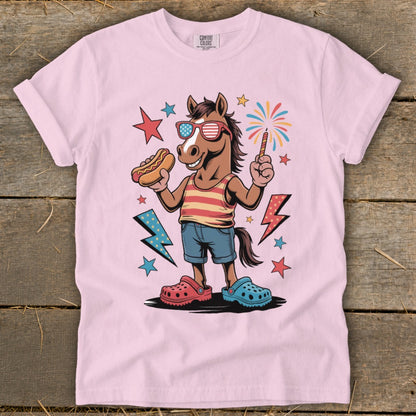 Cartoon horse wearing American flag sunglasses, holding hot dog and sparkler, on pink t-shirt.