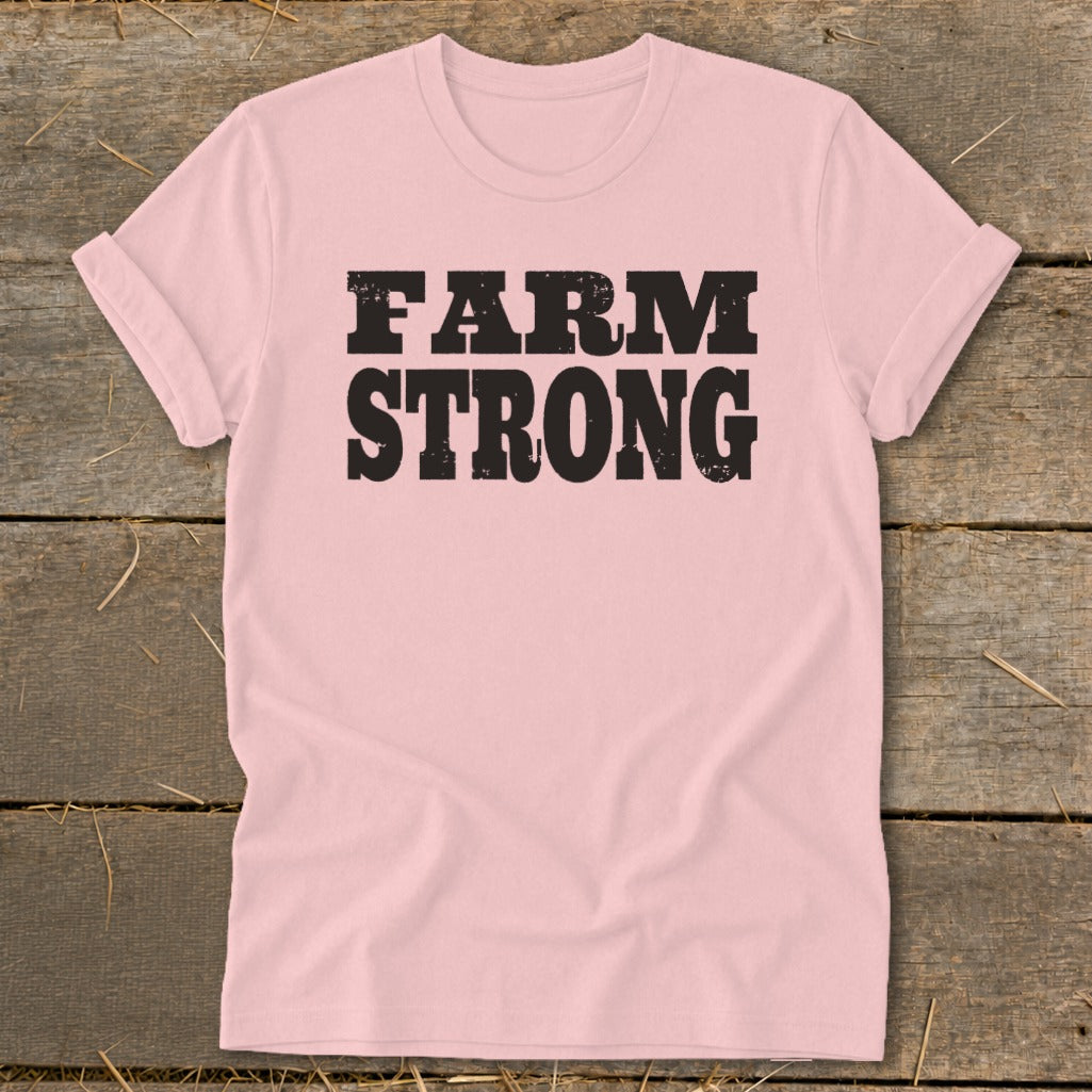 Pink t-shirt with distressed black text reading FARM STRONG, laid flat on a rustic wooden surface.