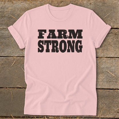 Pink t-shirt with distressed black text reading FARM STRONG, laid flat on a rustic wooden surface.