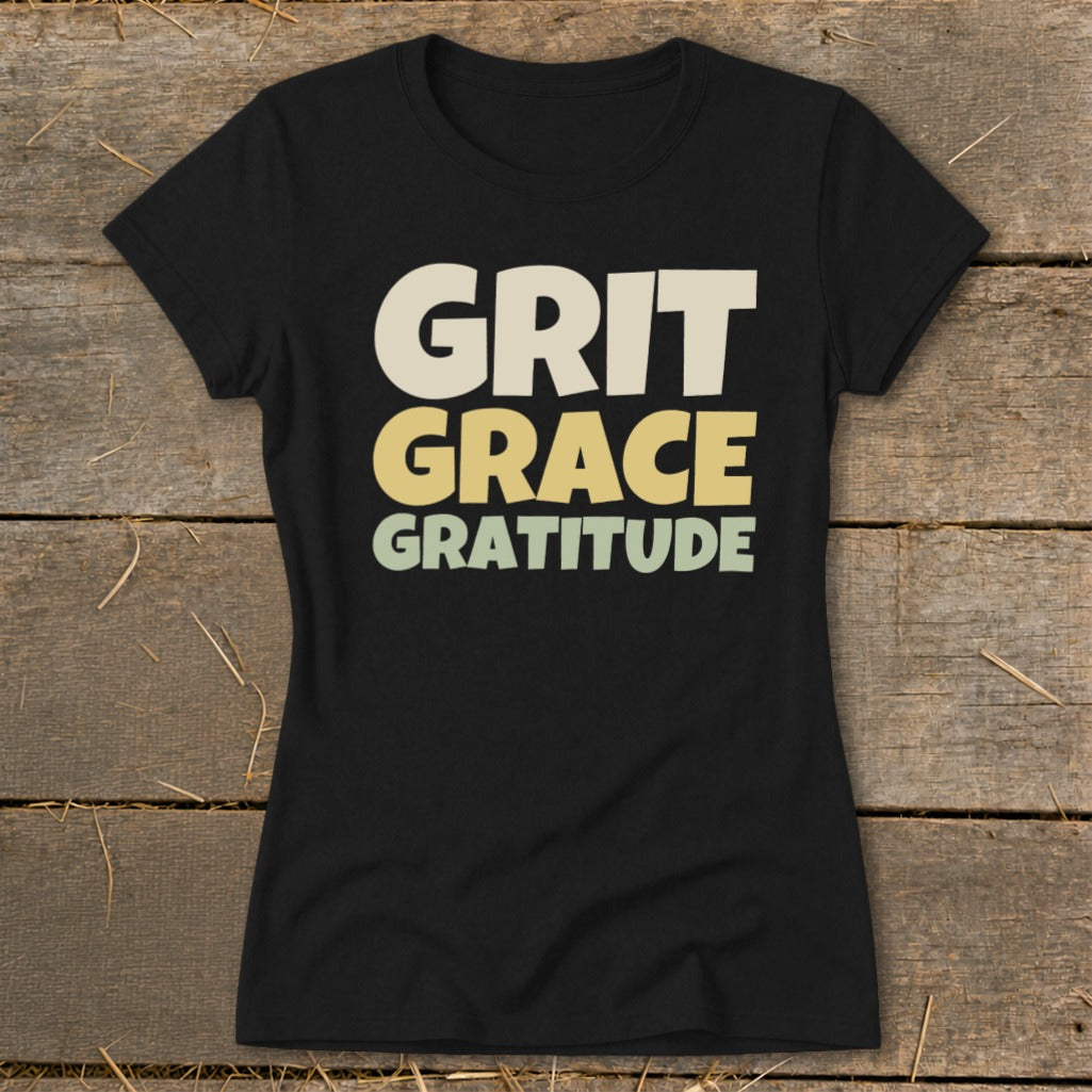 Black t-shirt displaying stacked retro text that reads Grit Grace Gratitude against a wooden background.