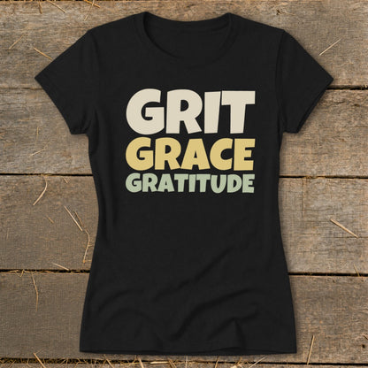 Black t-shirt displaying stacked retro text that reads Grit Grace Gratitude against a wooden background.