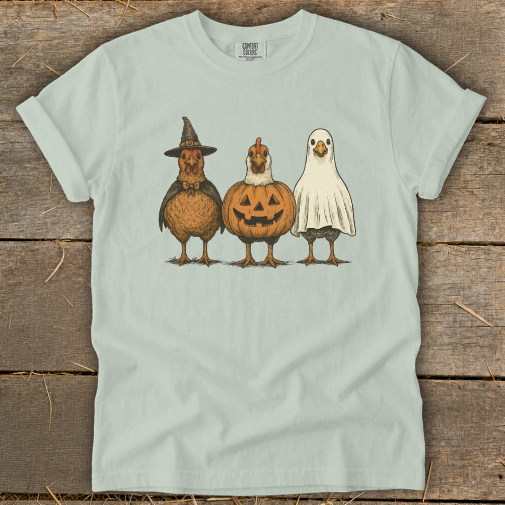 Three chickens dressed as a witch, pumpkin, and ghost on a light green t-shirt.