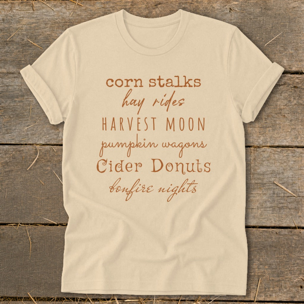 Light tan t-shirt with text: corn stalks, hay rides, Harvest Moon, pumpkin wagons, Cider Donuts, bonfire nights.