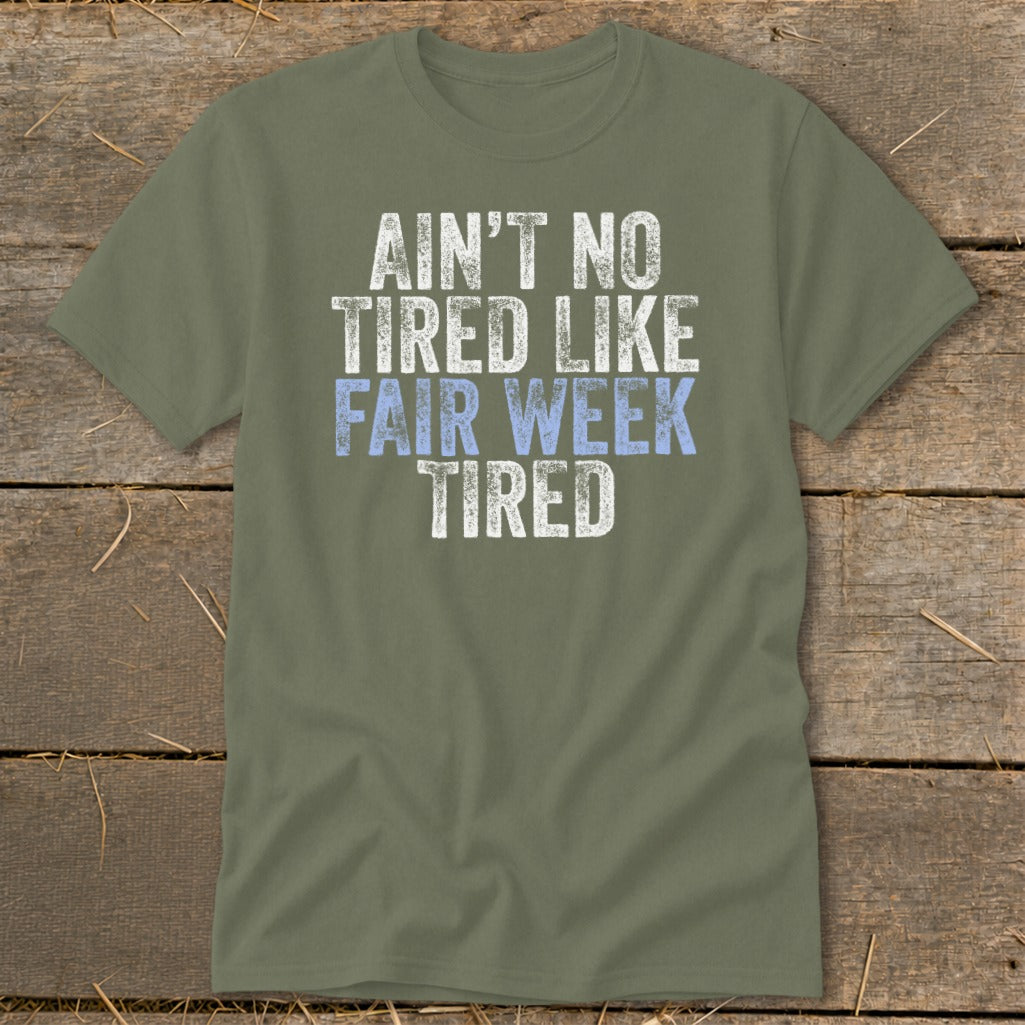 Olive green t-shirt with white and light blue text that says "Ain't No Tired Like Fair Week Tired".