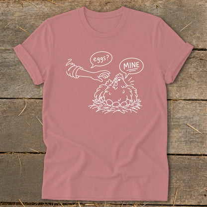 Pink t-shirt with white illustration of a chicken protecting eggs, saying "MINE".