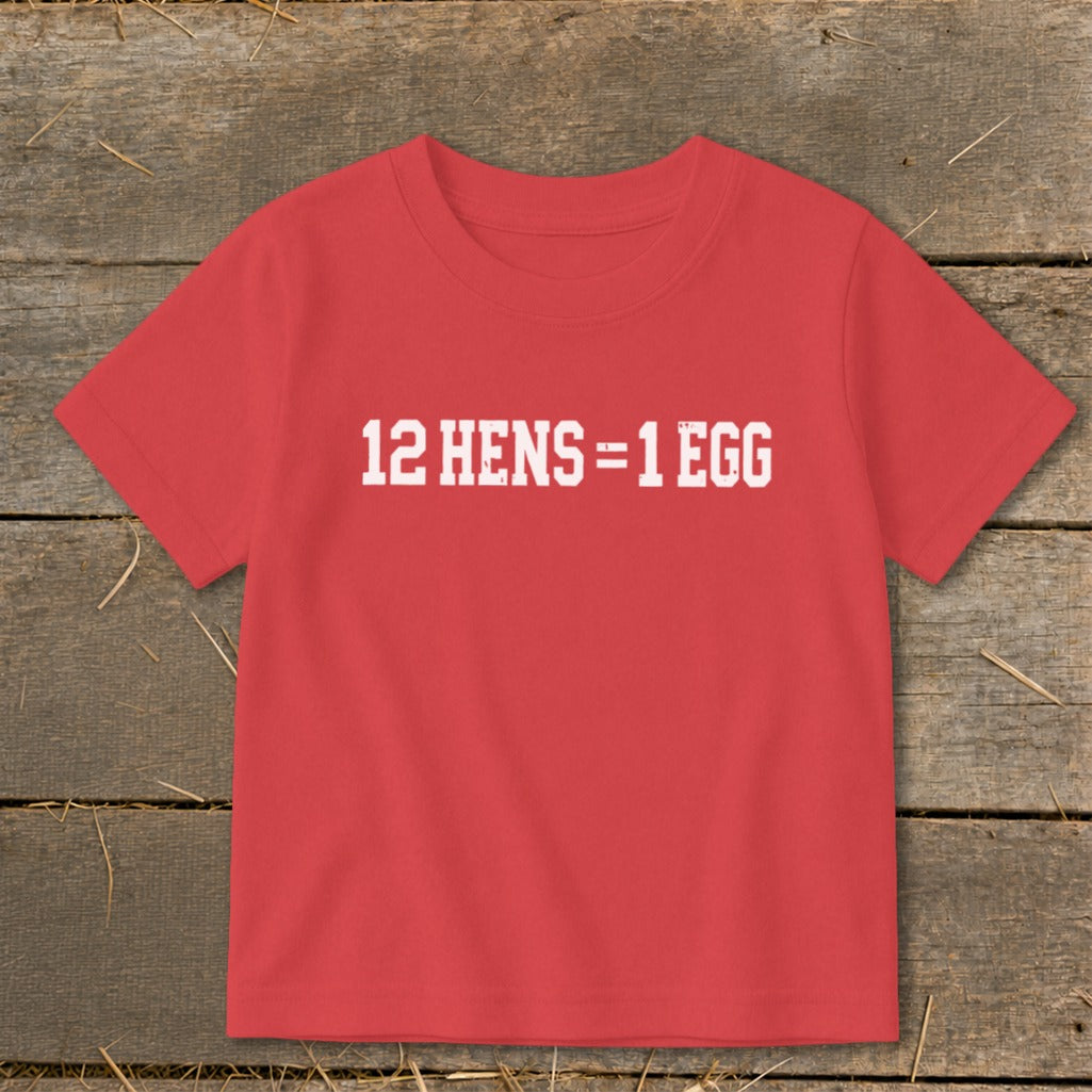 A red t-shirt with white distressed text reading 12 HENS = 1 EGG, laid flat on a rustic wooden background.