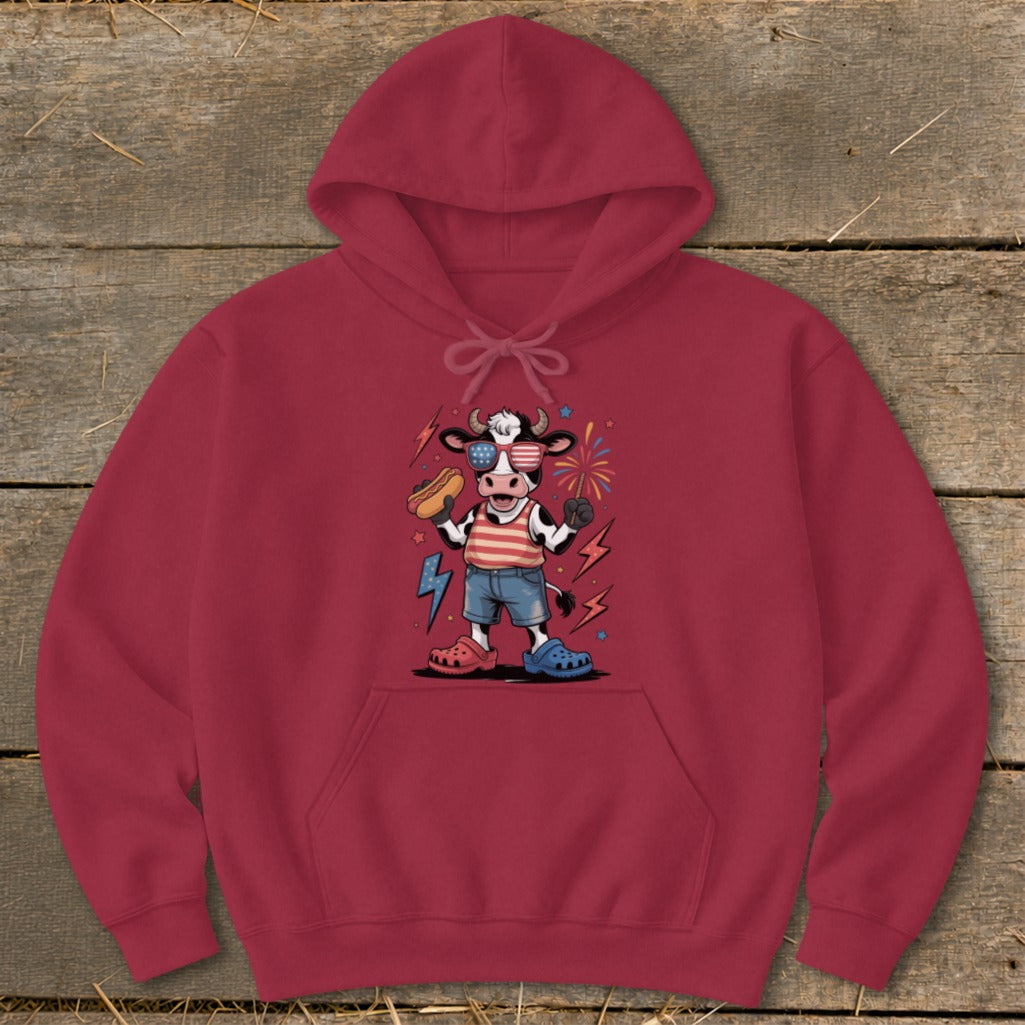 Cartoon cow wearing American flag clothes and sunglasses, holding a hot dog and sparkler.