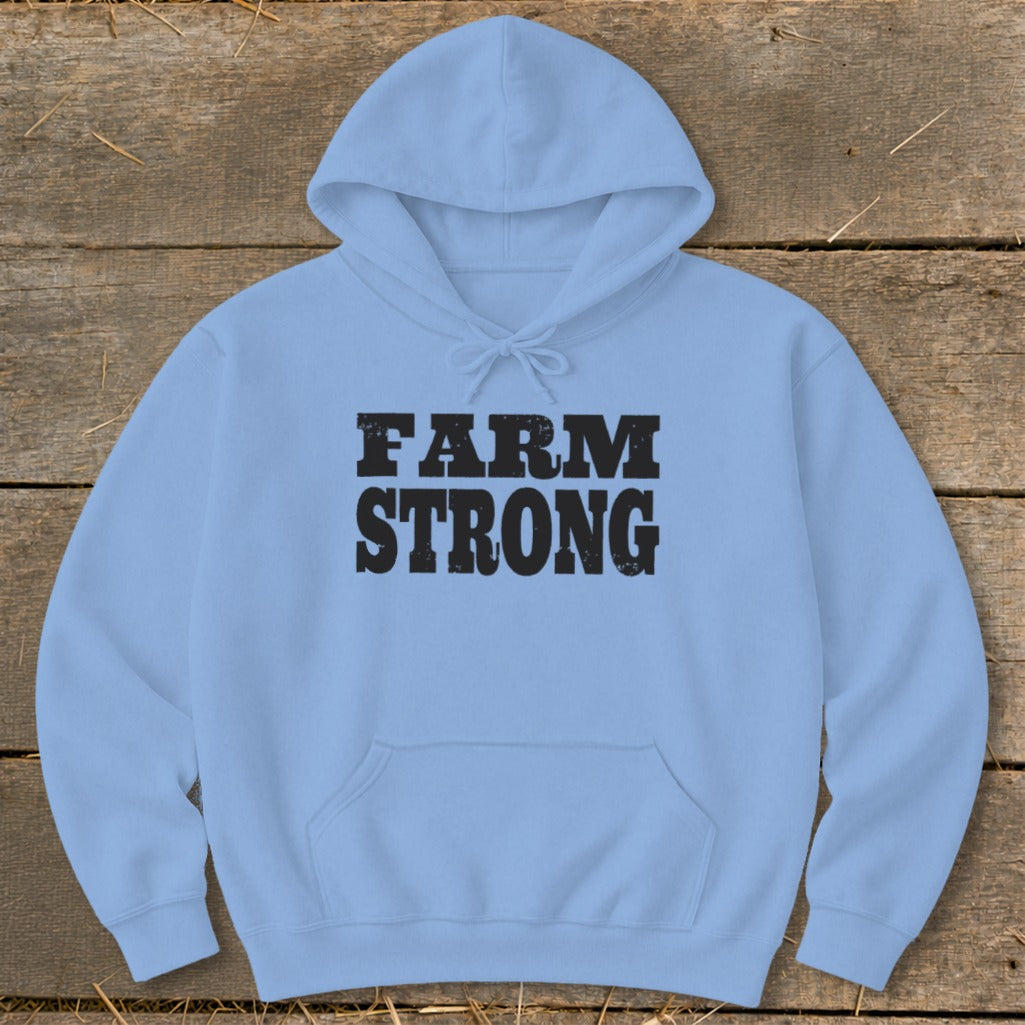 A light blue hooded sweatshirt featuring the black distressed text "FARM STRONG" across the chest, laid flat on a rustic wooden background.