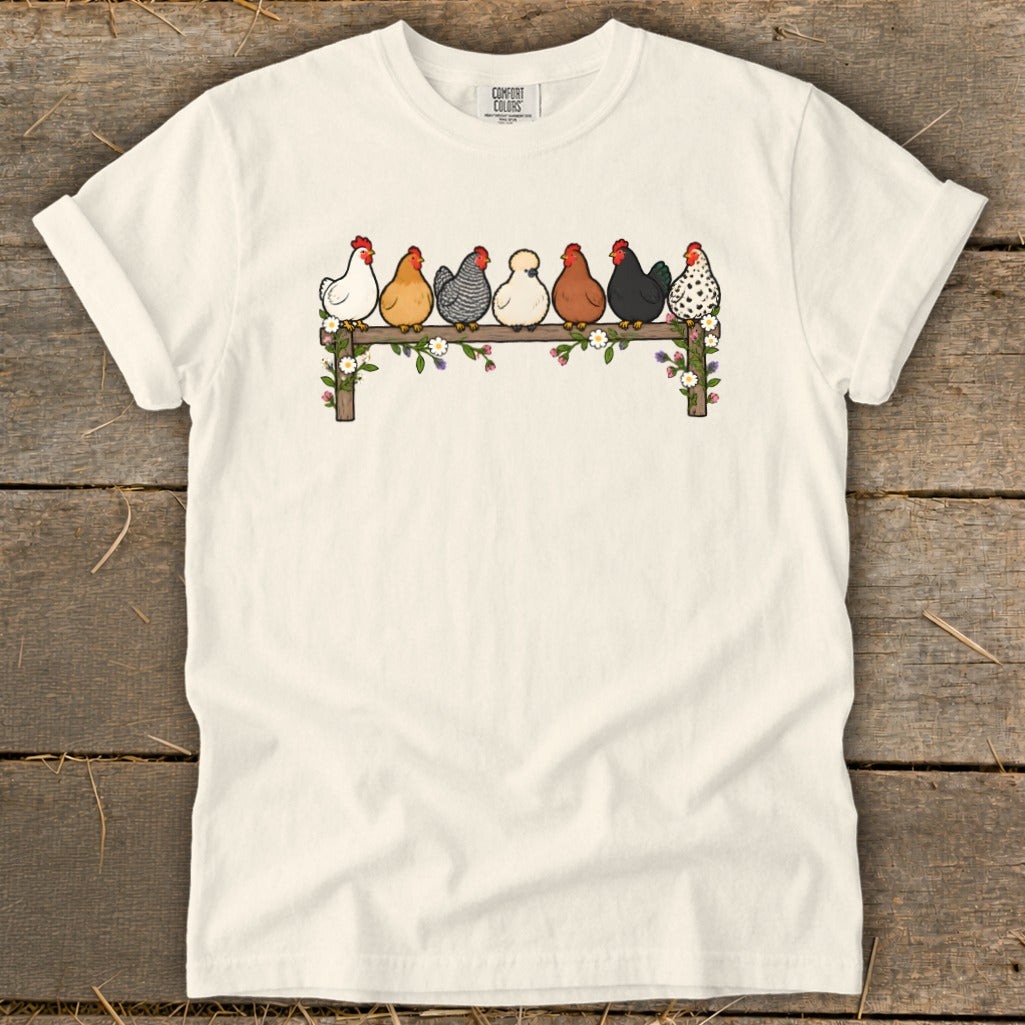 A cream-colored t-shirt with an illustration of seven different chickens perched on a flower-covered wooden fence.