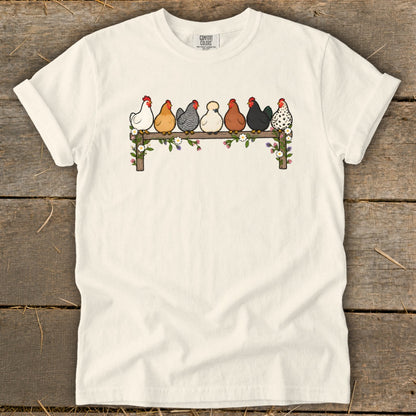 A cream-colored t-shirt with an illustration of seven different chickens perched on a flower-covered wooden fence.