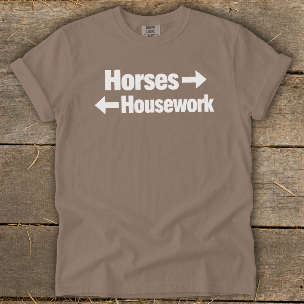 A brown t-shirt featuring white text that reads Horses with a right arrow above Housework with a left arrow.