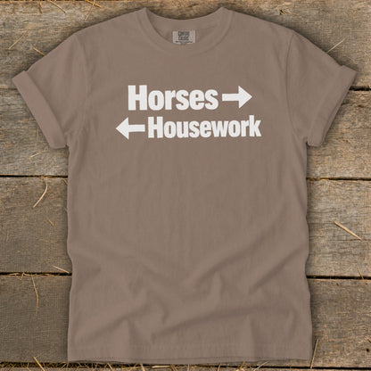 A brown t-shirt featuring white text that reads Horses with a right arrow above Housework with a left arrow.