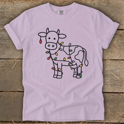 Light purple t-shirt with a black outline drawing of a cow wrapped in colorful Christmas lights.