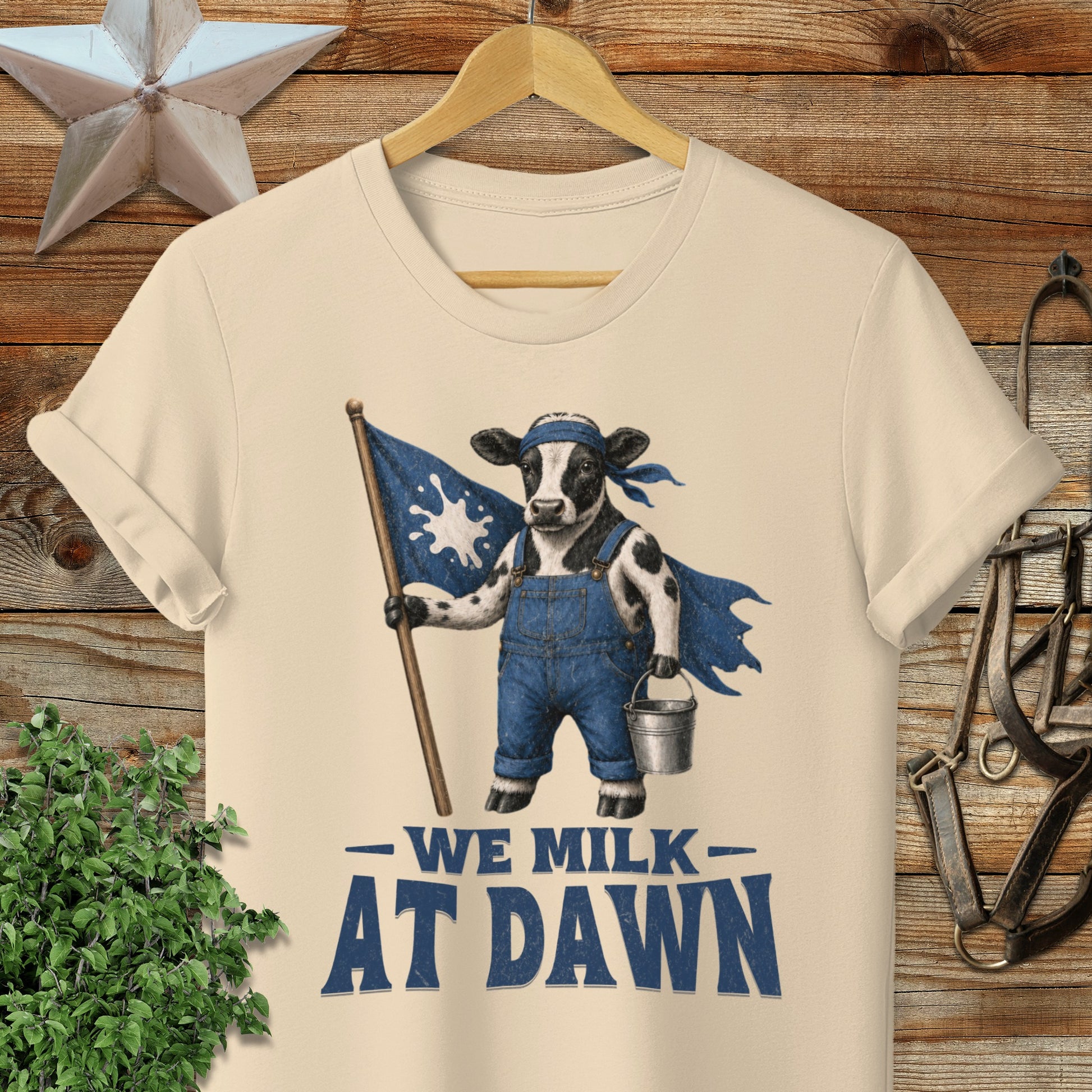 Cow in overalls holding a flag and milk bucket with text "We Milk At Dawn".