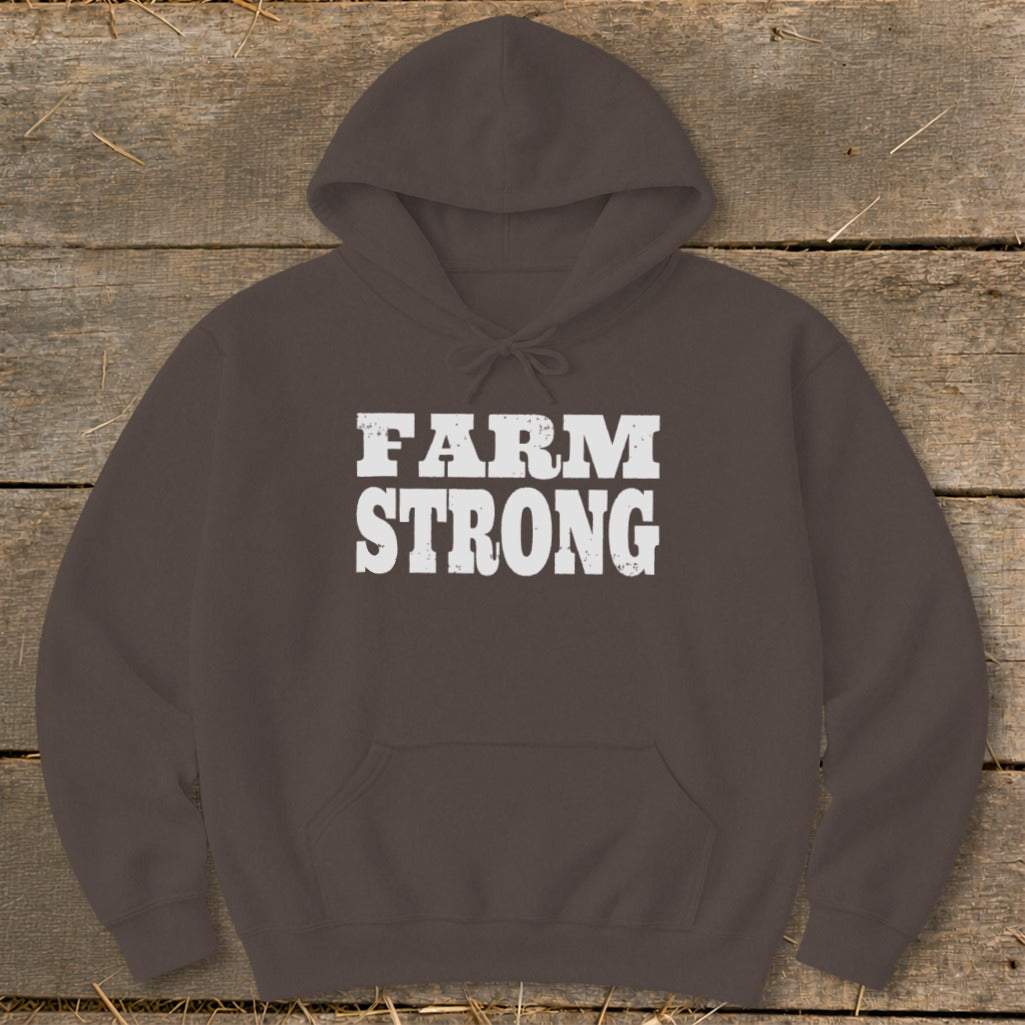 Dark brown hooded sweatshirt with the white distressed text "FARM STRONG" centered on the chest, lying flat on a rustic wooden background.