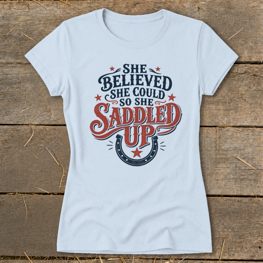Light blue t-shirt with "She believed she could so she saddled up" text and horseshoe design.